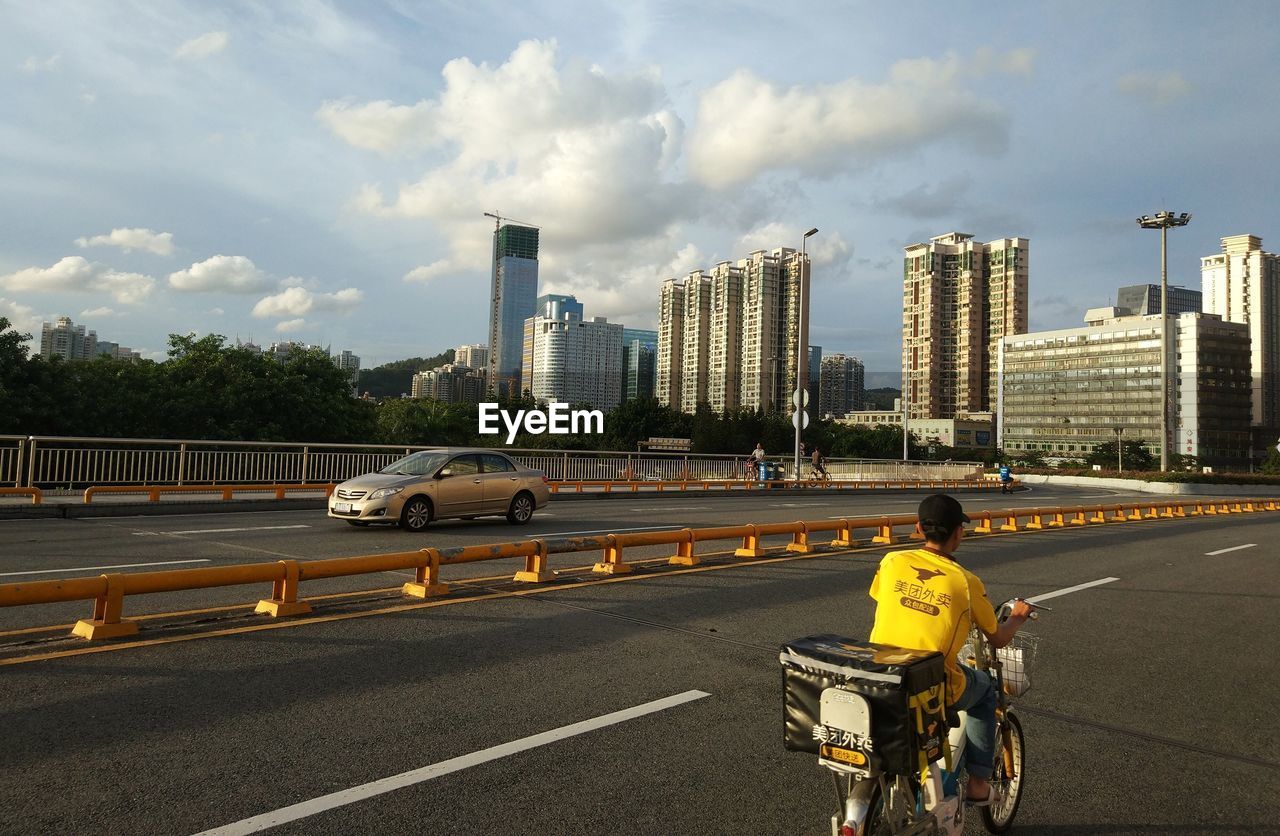 REAR VIEW OF MAN CYCLING ON ROAD AGAINST SKY