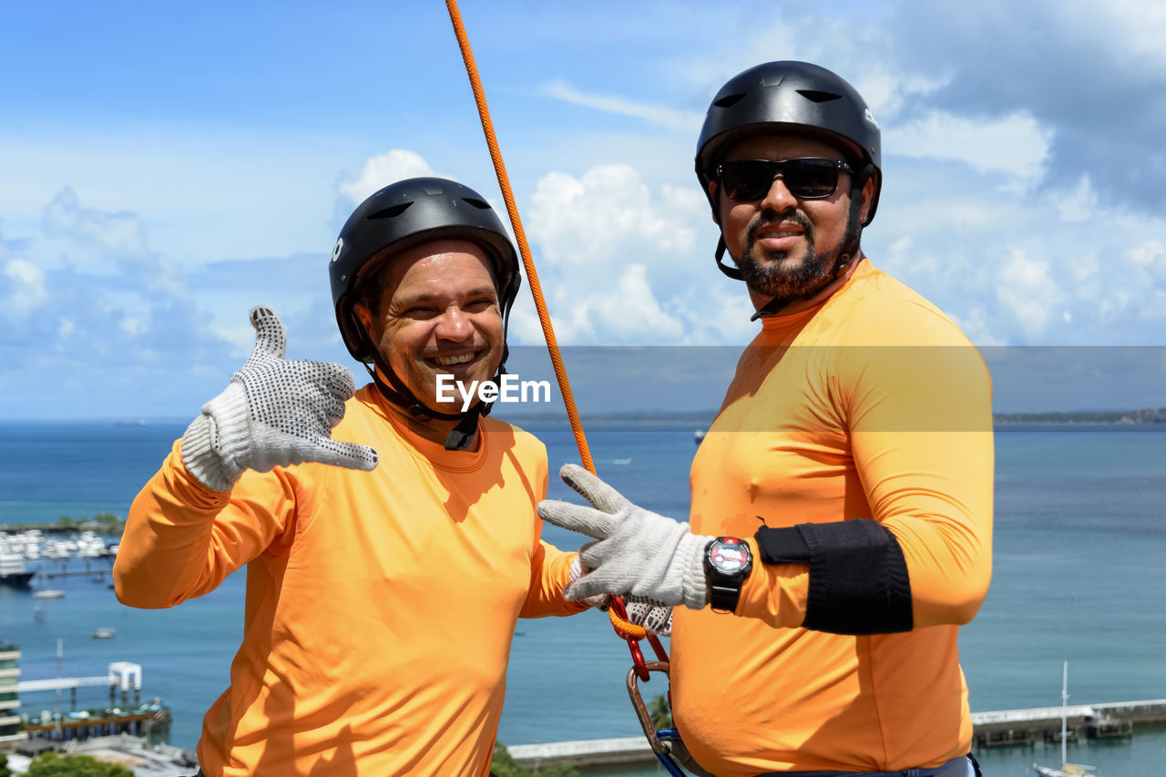 Portrait of two smiling men standing holding a rappel rope against the sky.
