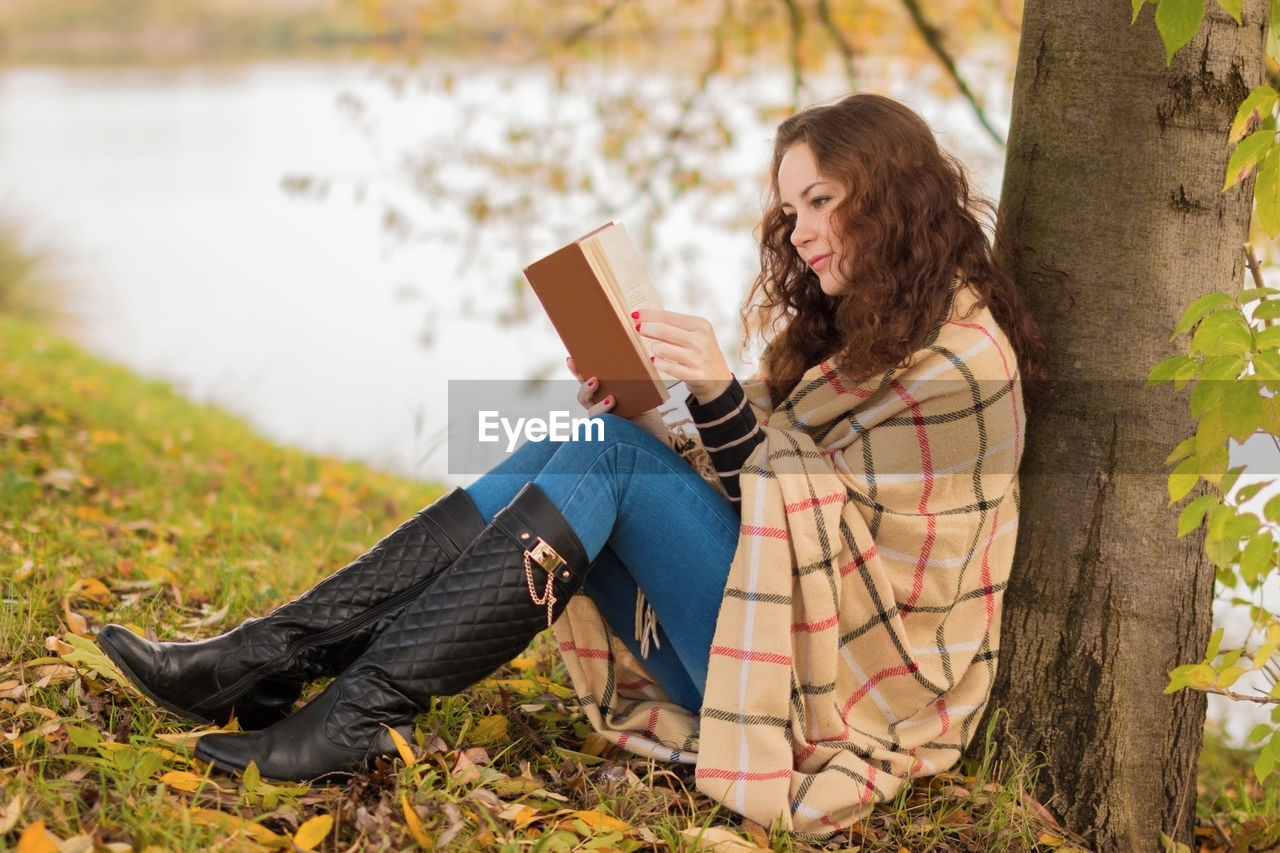 Side view of woman reading book while leaning on tree trunk at lakeshore