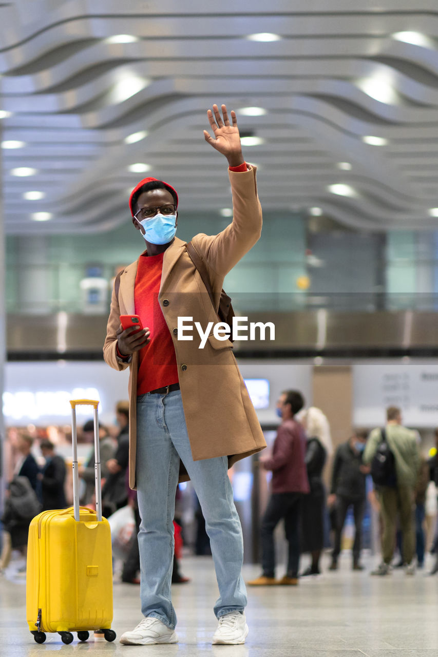 Full length of man wearing mask standing with hand raised at airport