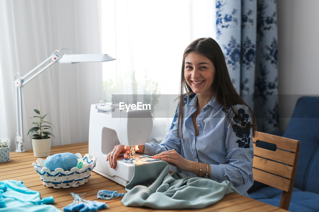 adult, women, smiling, one person, technology, indoors, sitting, happiness, communication, wireless technology, table, computer, business, furniture, working, young adult, lifestyles, business finance and industry, emotion, female, home interior, businesswoman, hairstyle, person, office, casual clothing, occupation, cheerful, portrait, long hair, domestic room, laptop, internet, digital tablet, computer network, desk, brown hair, clothing, domestic life, looking at camera, businesswear, black hair, smile, teeth, relaxation, portability, using laptop, holding, small business, entrepreneur, enjoyment, using computer, looking, living room, front view, convenience