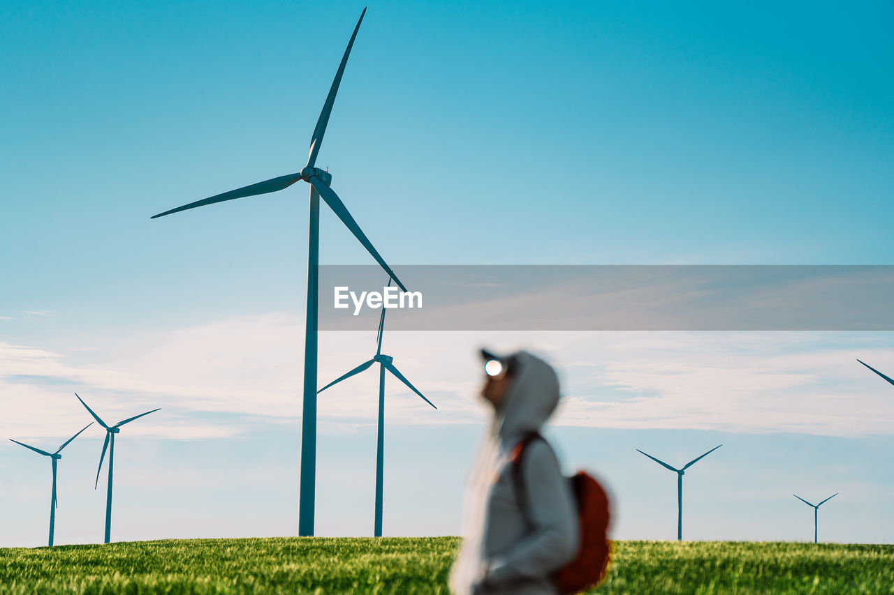 Person standing by wind turbine in agricultural field against clear blue sky