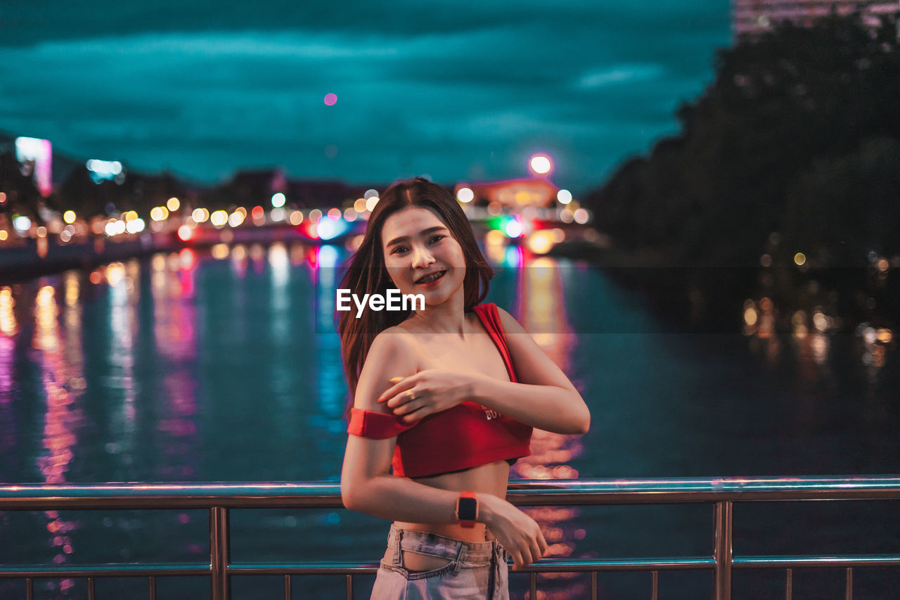 Portrait of smiling young woman standing on bridge over river at night