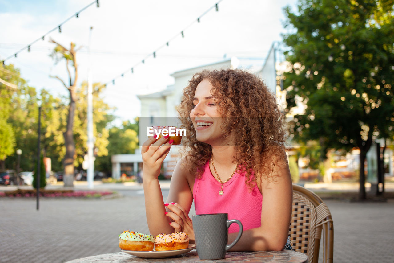 Young attractive curly woman sits at table in cafe on summer terrace. colorful delicious donuts, 