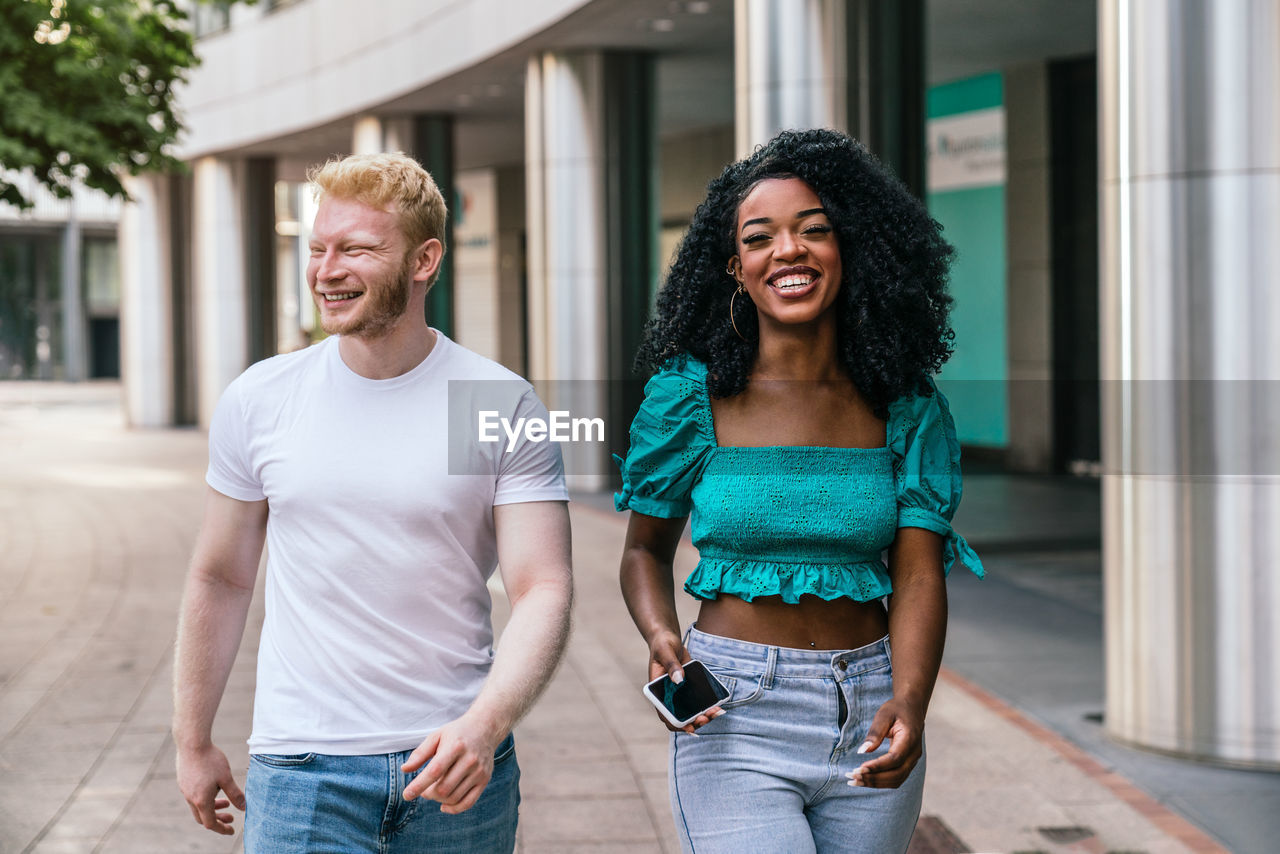 Smiling african american woman in blue top and jeans with afro hairstyle and blond haired man in white t shirt walking along paved sidewalk and chatting while looking at camera