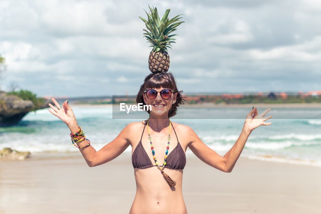 Portrait of happy young woman in bikini balancing pineapple on head at beach
