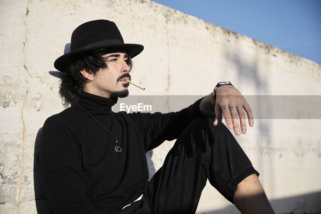 Side view of young bearded guy in stylish black clothes and hat smoking cigarette and looking away thoughtfully while sitting near shabby concrete wall on street