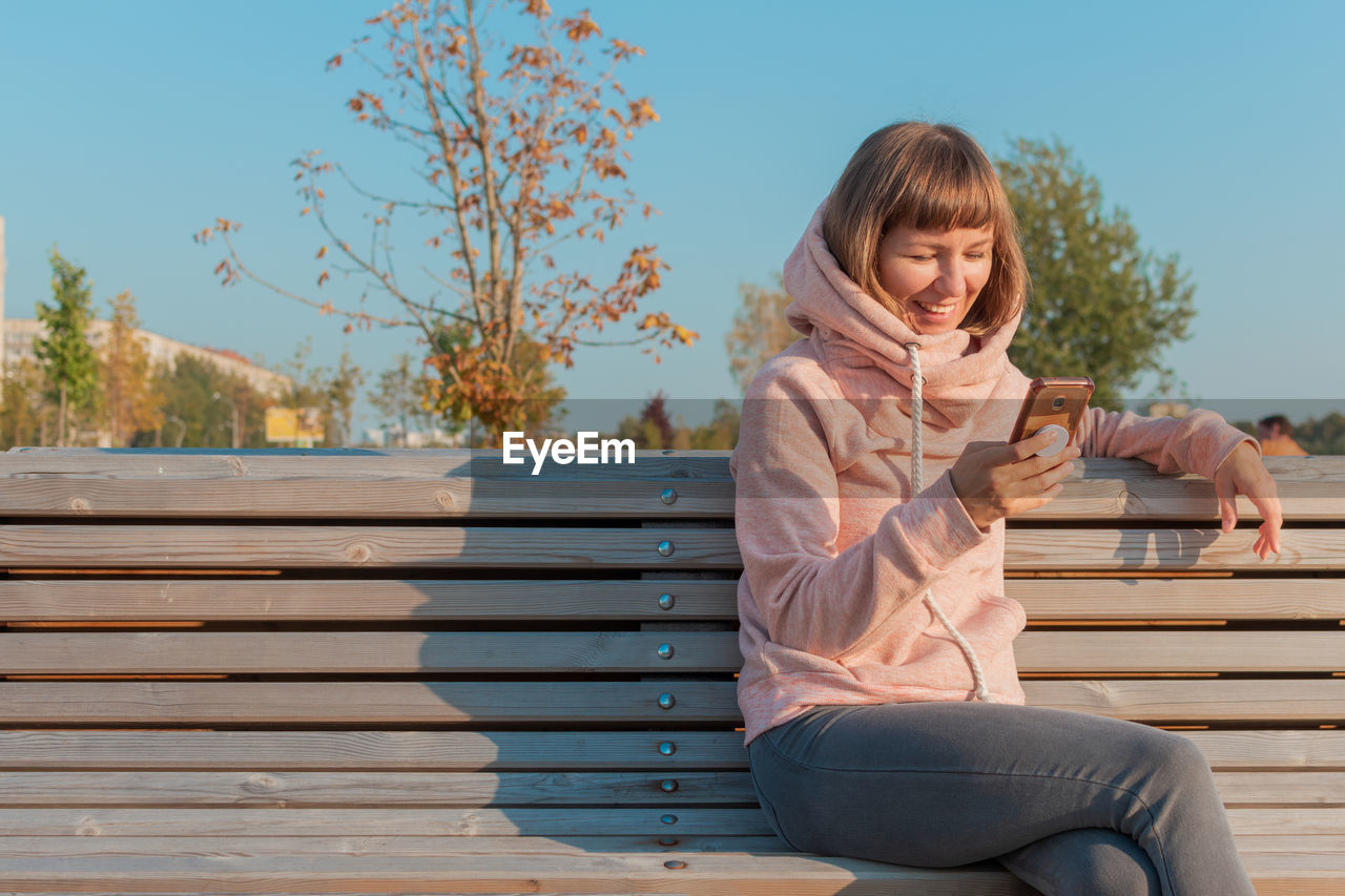 Smiling young woman using mobile phone while sitting on bench