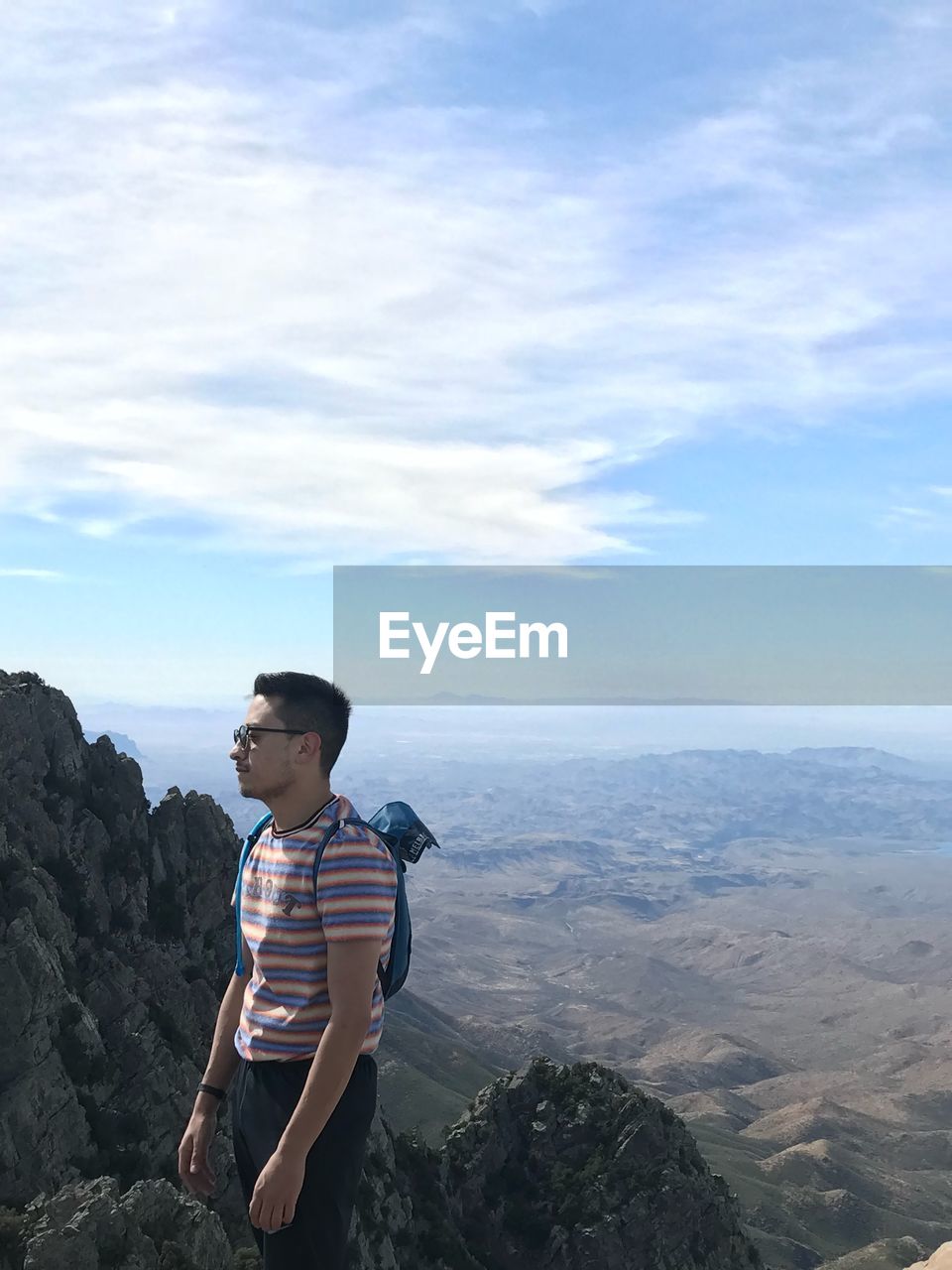 Side view of young man standing on mountain peak against cloudy sky