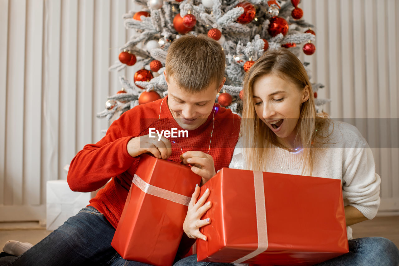 Young couple sitting near christmas tree and starting unpack presents together with excitement