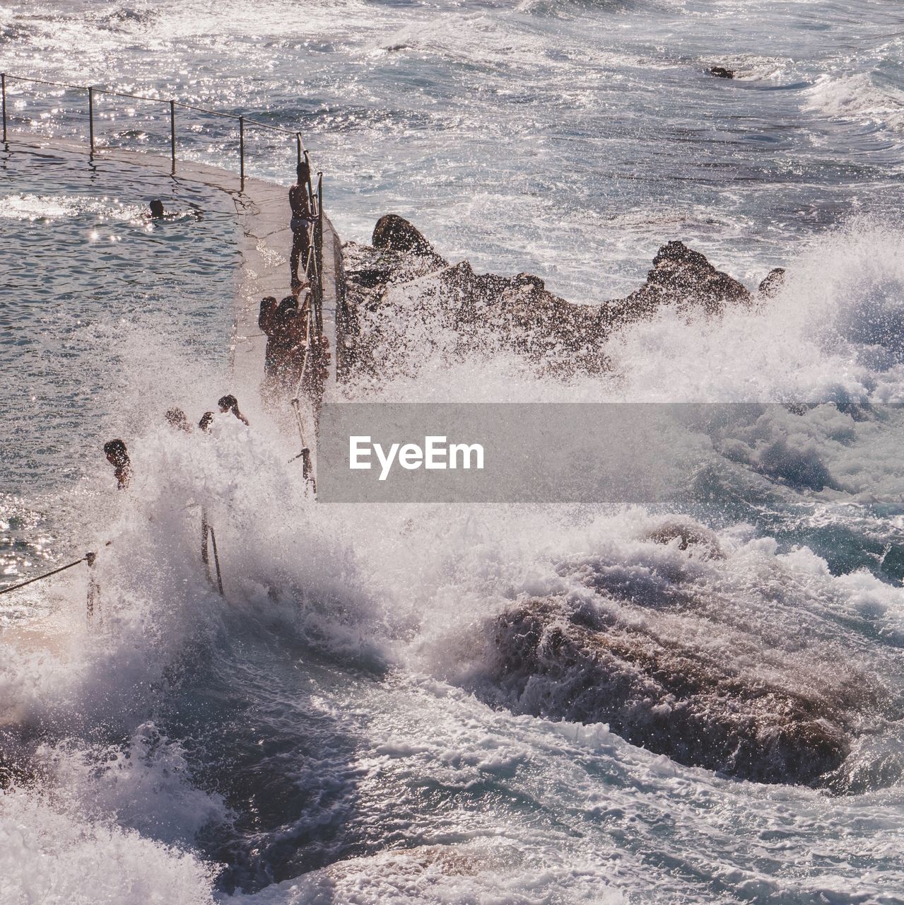 High angle view of waves splashing on people standing on retaining wall with fence