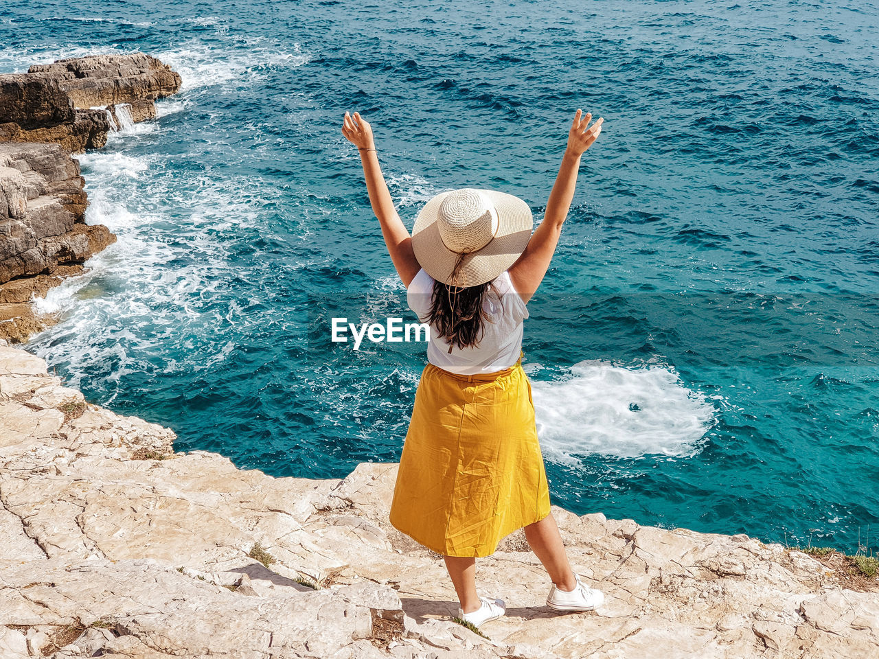 Young woman in yellow skirt standing on edge of cliff above sea.