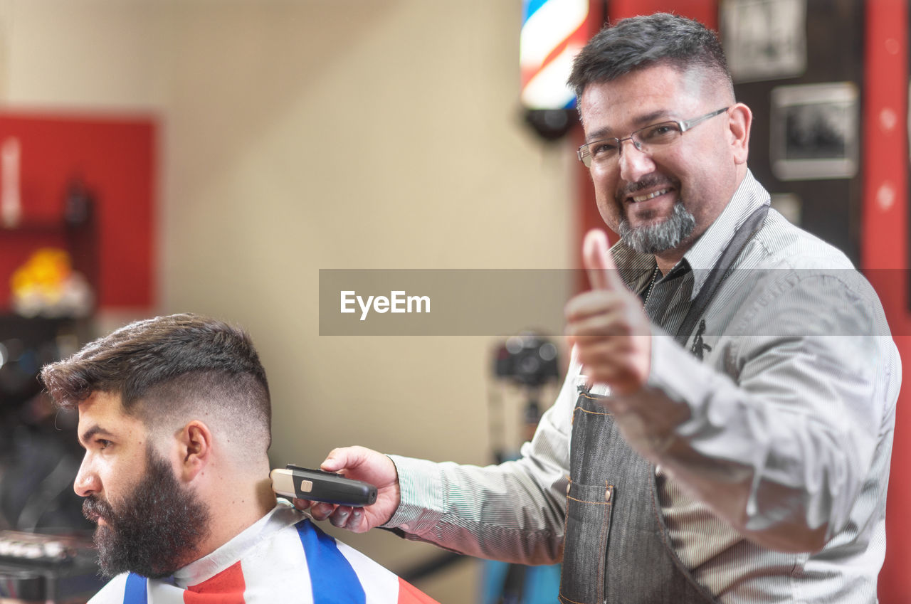 Portrait of smiling barber showing thumbs up while cutting male customer hair in salon