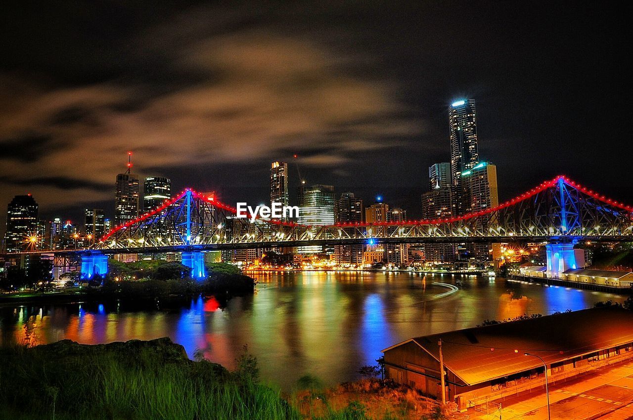 Illuminated bridge over river against buildings at night