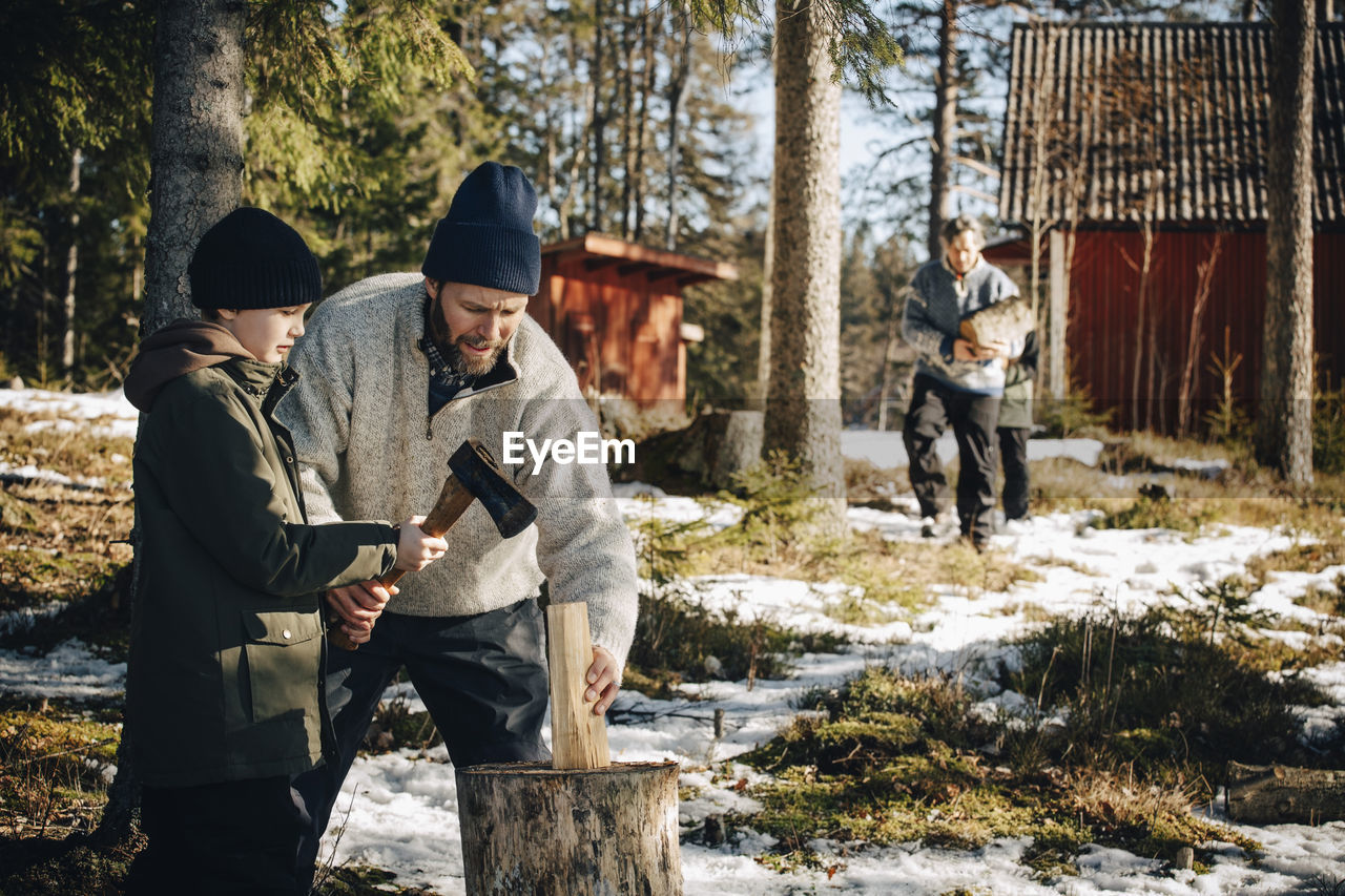 Father teaching son to cut log with axe in forest during winter