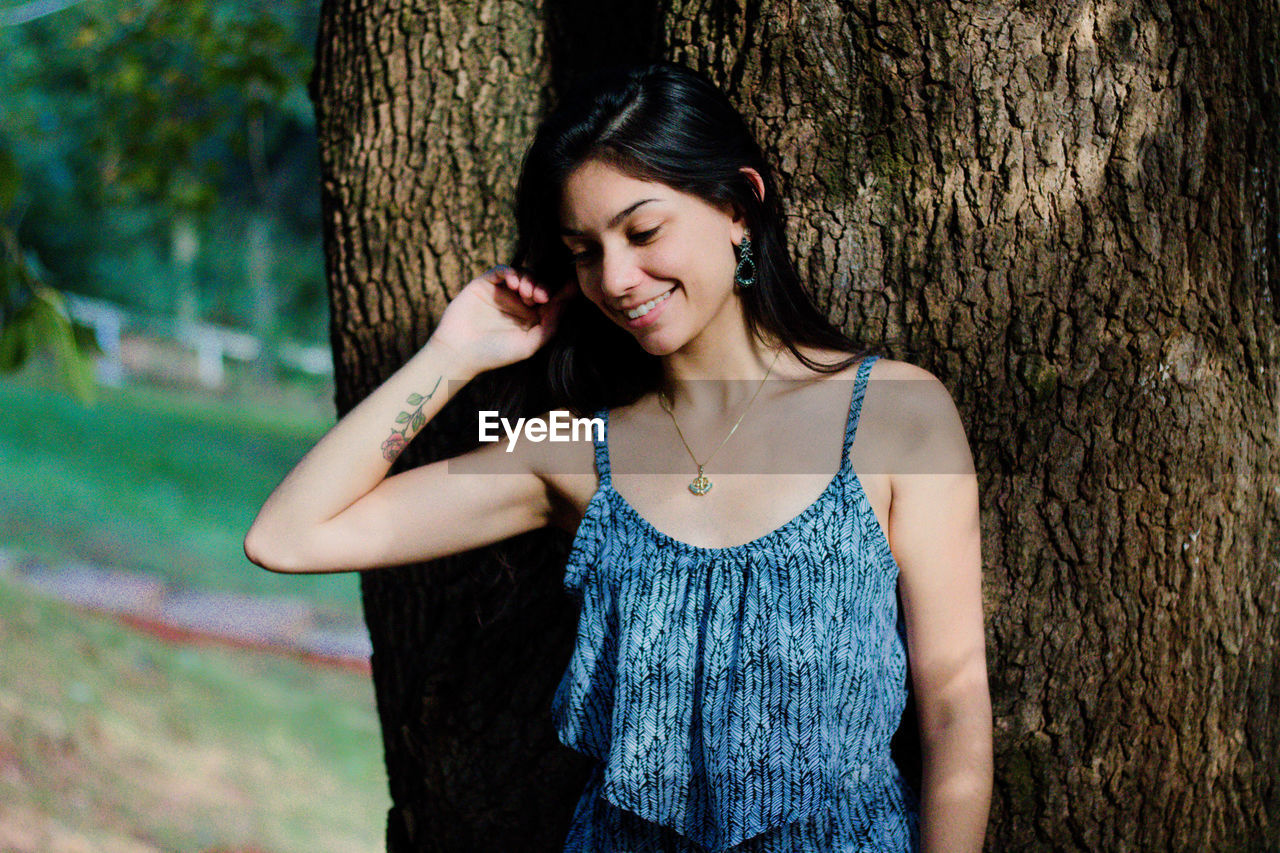 Beautiful young woman standing by tree trunk at park