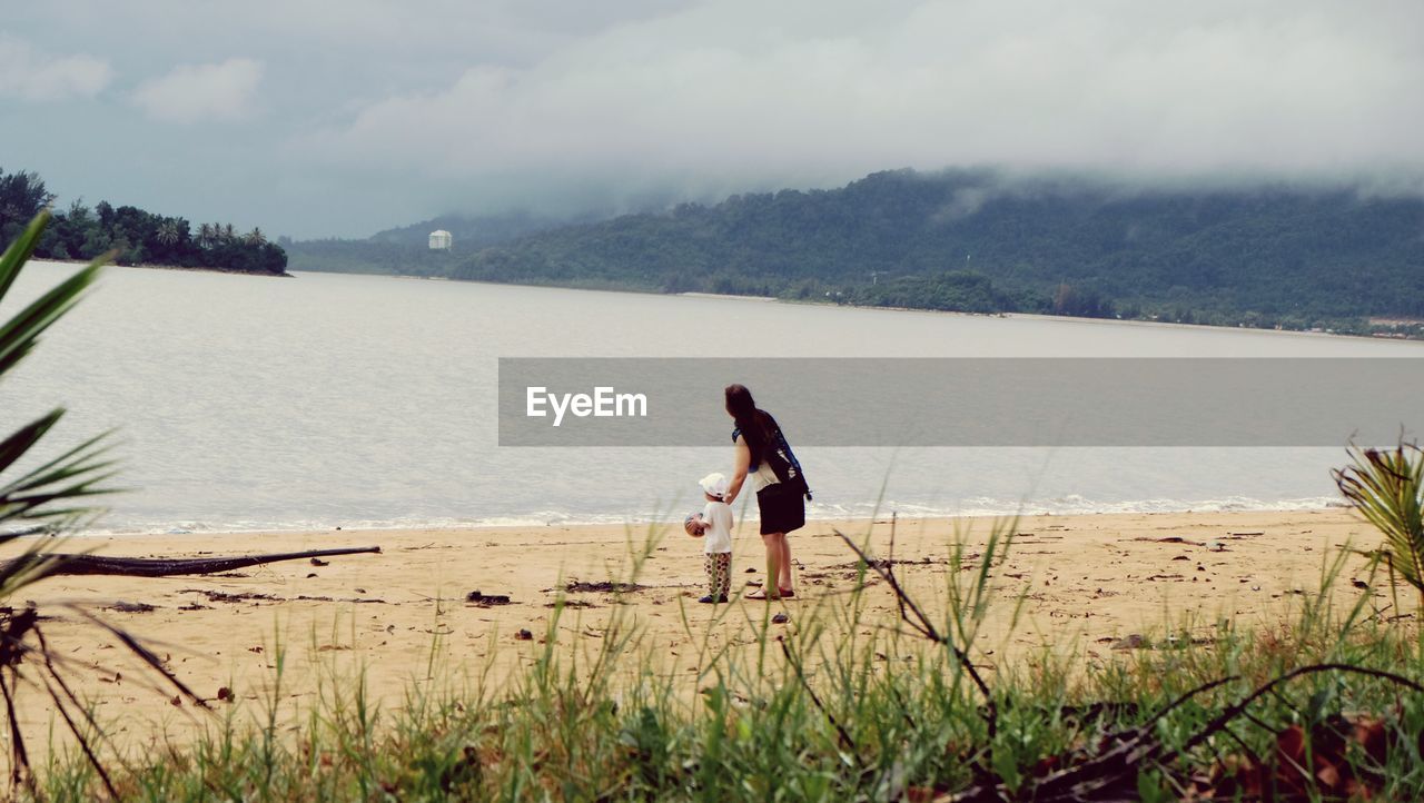 REAR VIEW OF SIBLINGS STANDING ON BEACH