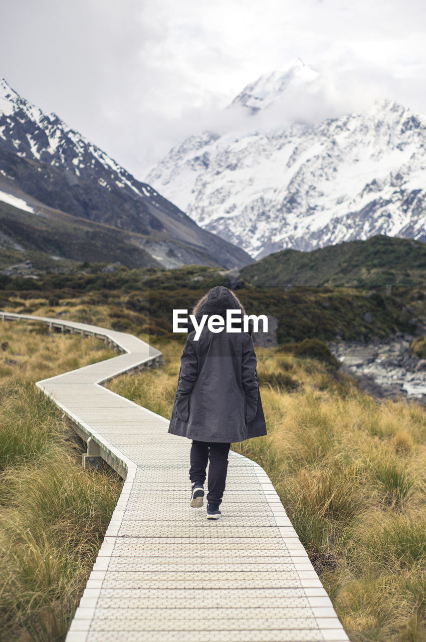 Rear view of woman wearing warm clothing while walking on boardwalk against snowcapped mountains