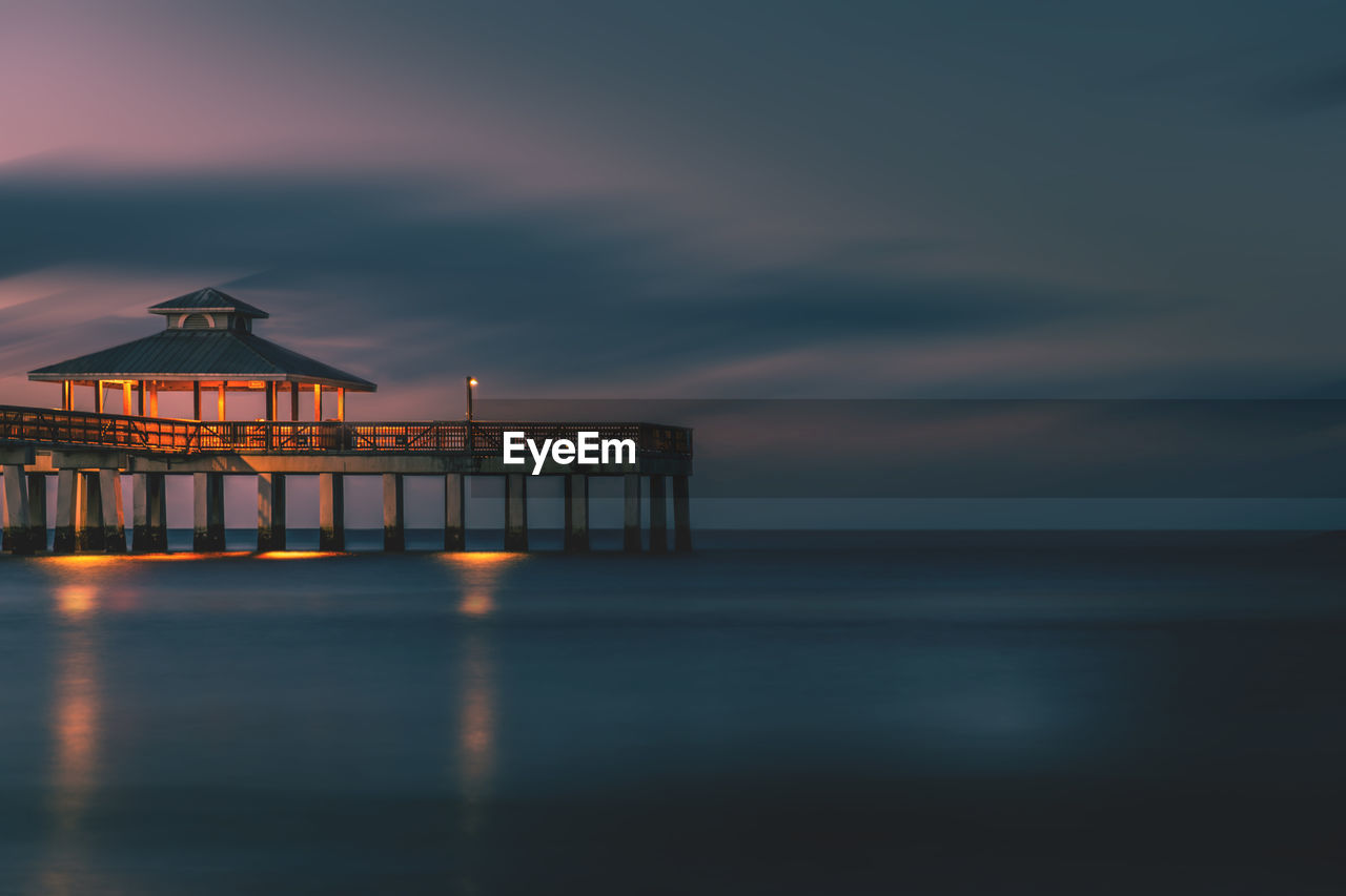 Illuminated built structure over sea against cloudy sky at dusk