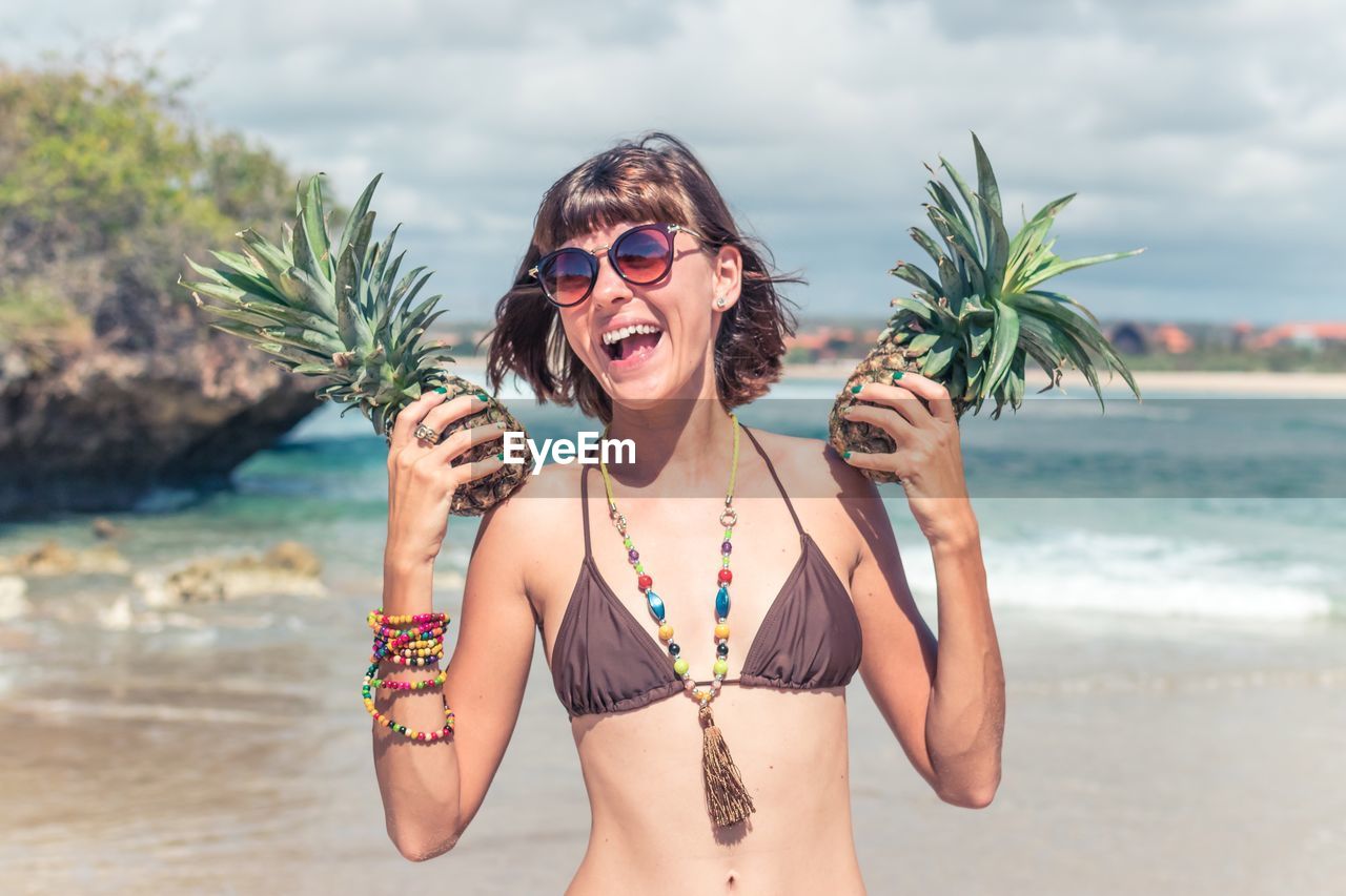 Happy young woman holding pineapples at beach against sky