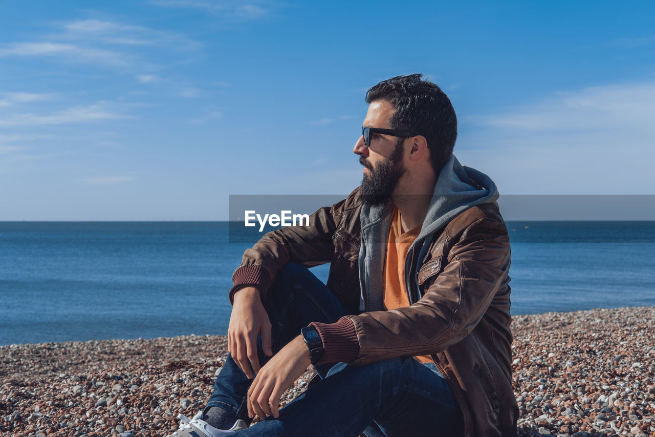 Side view of pensive ethnic bearded male traveler in trendy clothes and sunglasses relaxing on rocky beach of sea in seven sisters