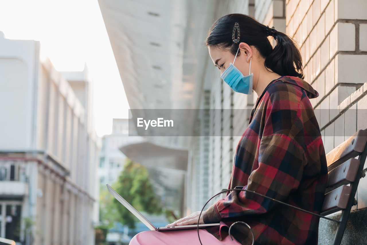 Woman wearing mask using laptop while sitting on bench outdoors