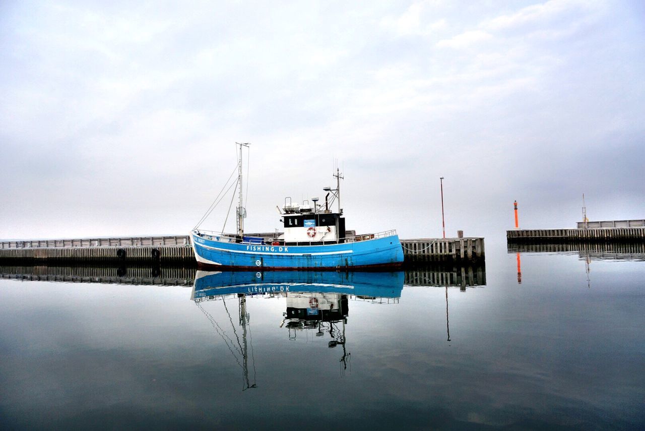 Trawler moored by piers over sea against cloudy sky