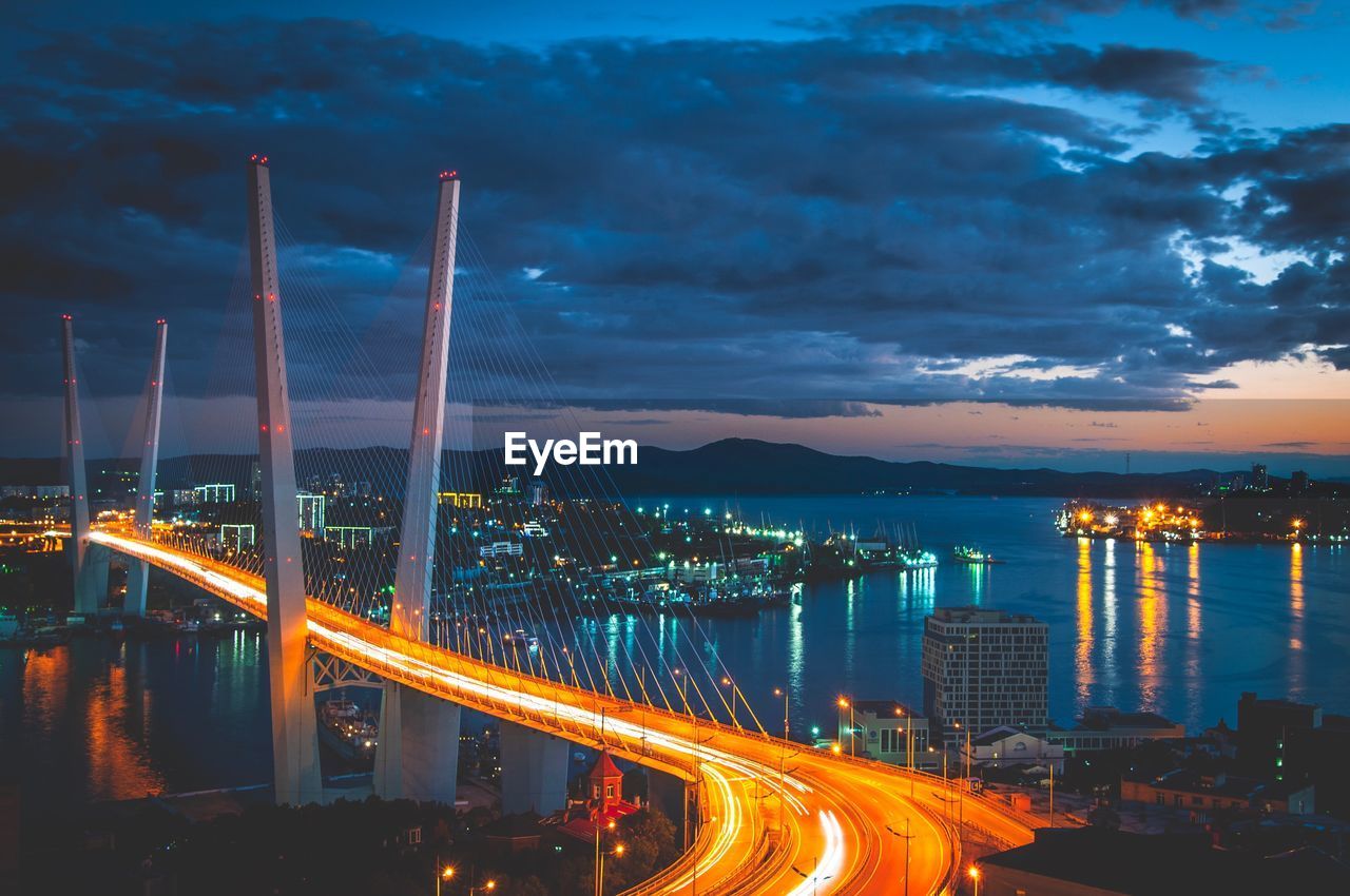 Light trails on bridge in city against cloudy sky at night