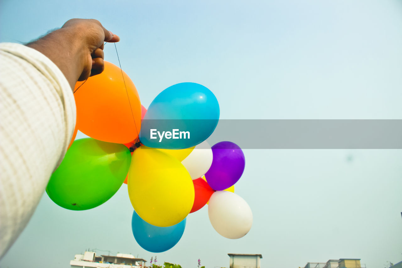 Cropped hand of man holding multi colored balloons against sky