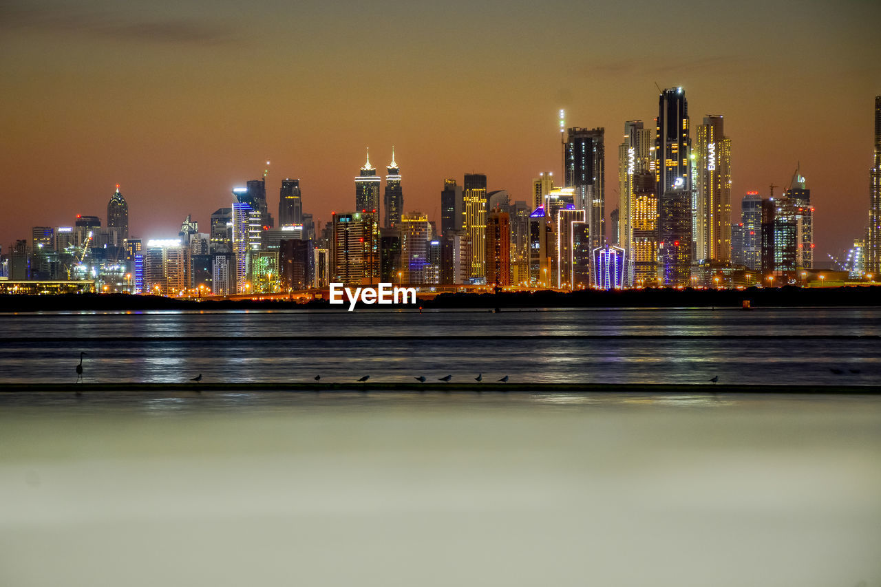 ILLUMINATED MODERN BUILDINGS BY RIVER AGAINST SKY AT NIGHT