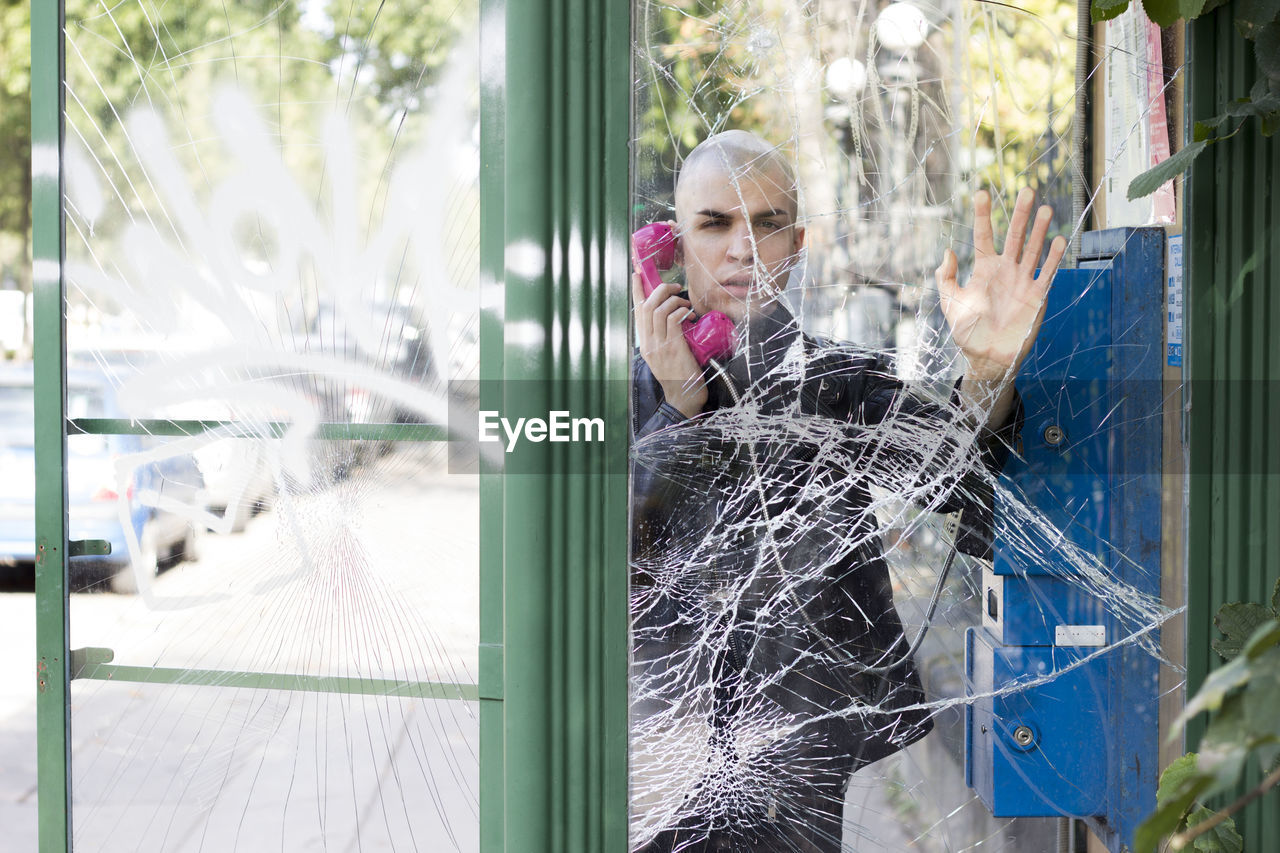 Portrait of bald man using telephone booth seen through shattered glass9