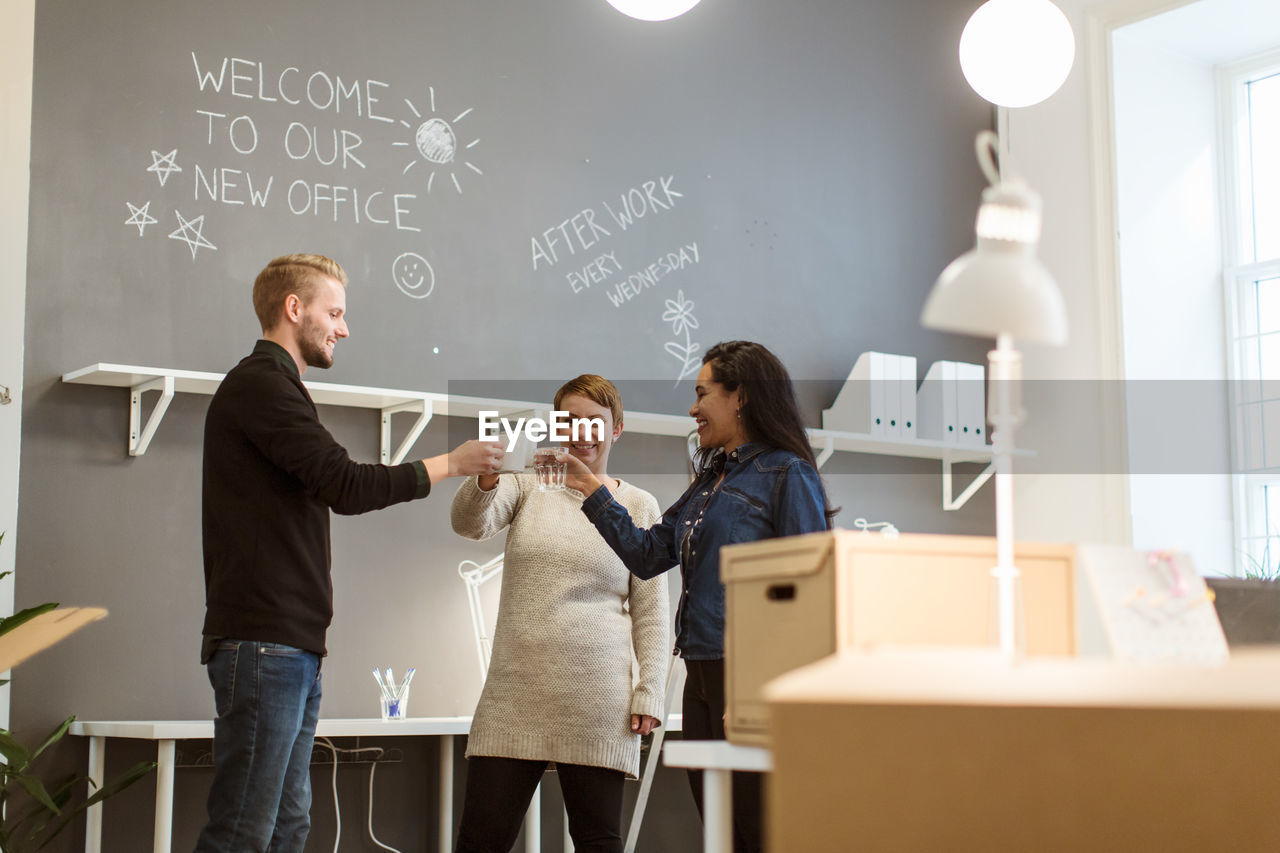 Low angle view of happy multi-ethnic colleagues toasting drinks while standing against wall at creative office