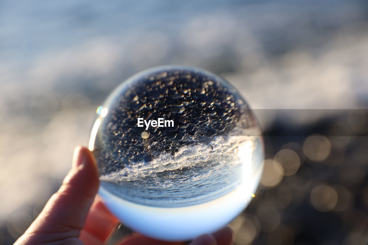 Cropped hand holding crystal ball against sea during sunny day