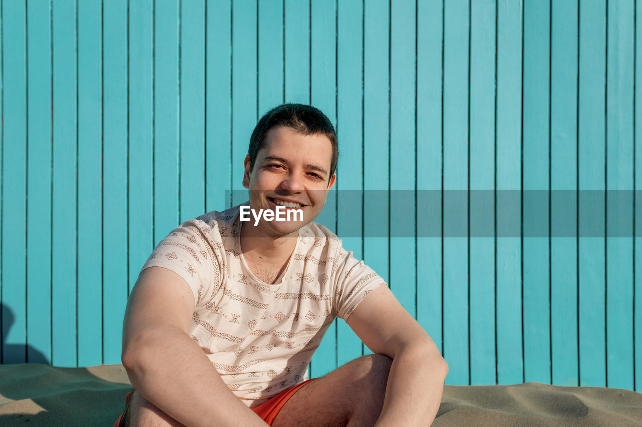 Young man sitting in sand beach smiling at camera in a turquoise wall