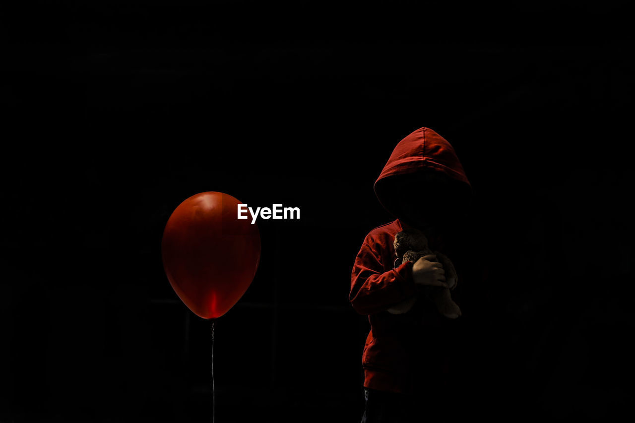 Boy wearing hood holding stuffed toy while standing by red balloon against black background