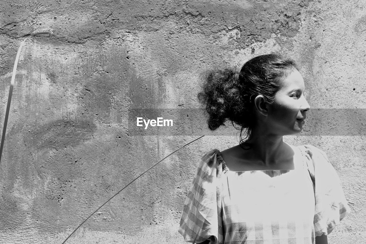 Close-up of young woman looking away while standing by wall on sunny day