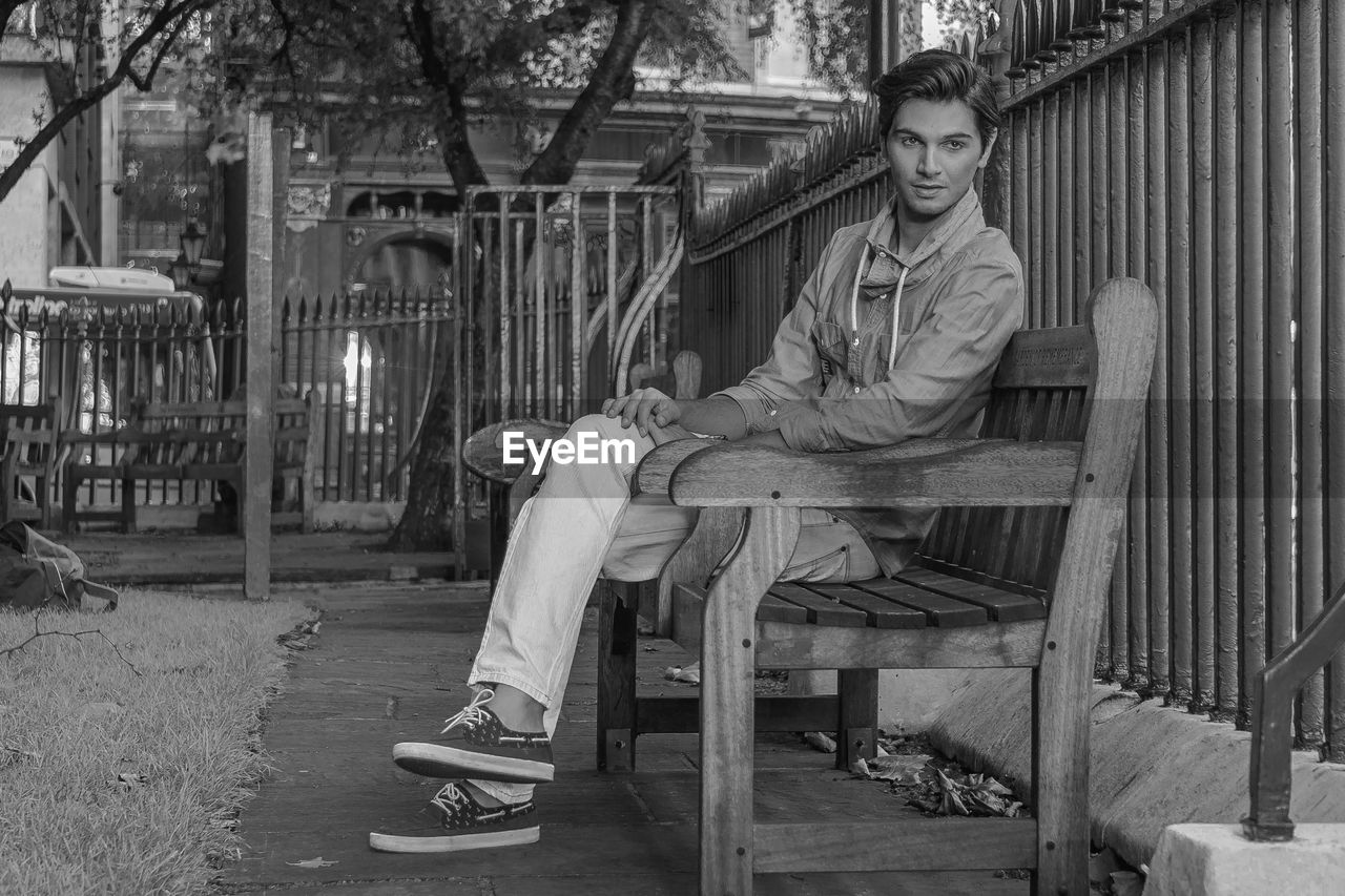 Full length of thoughtful young man sitting on bench by fence at park