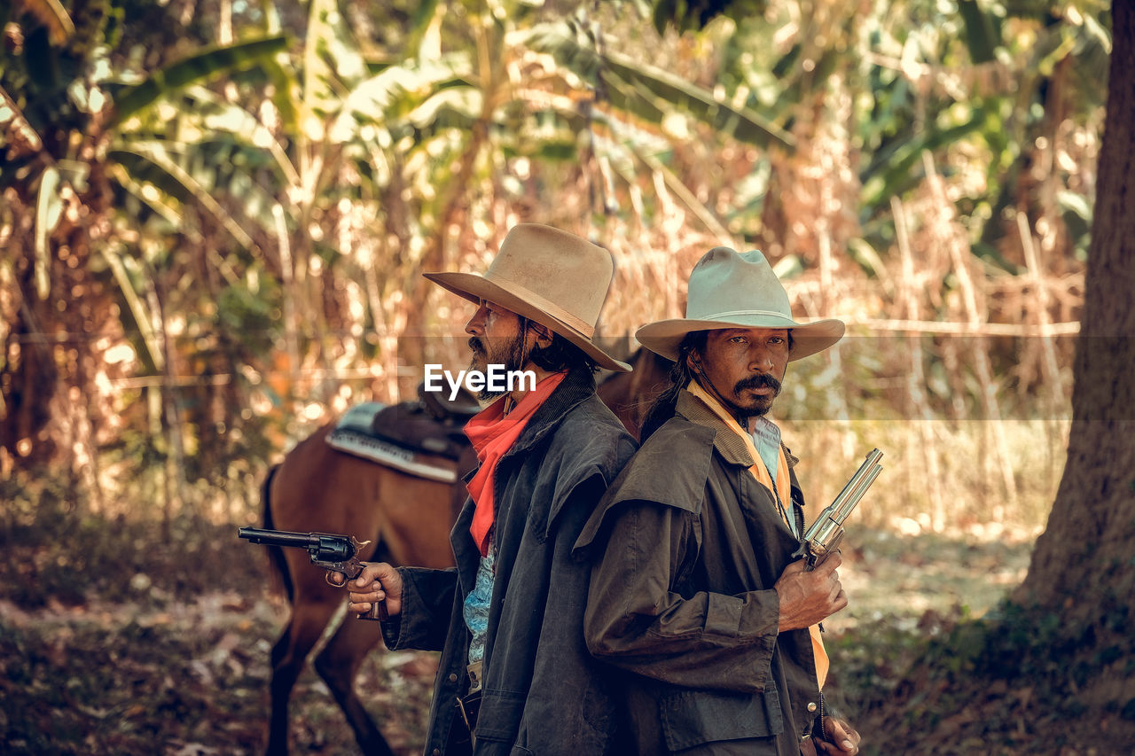 Men holding gun wearing hat standing with horse at forest