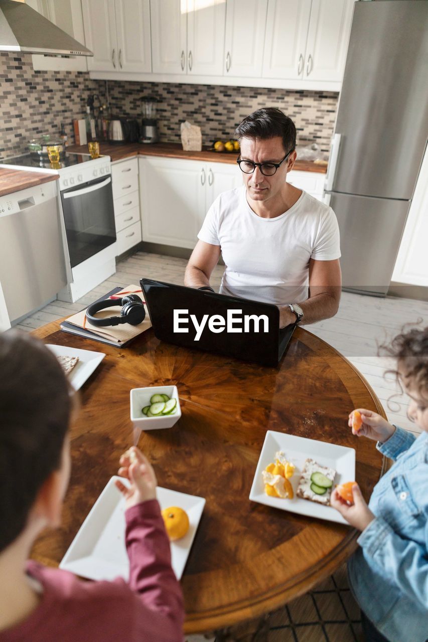 High angle view of siblings eating orange fruit while father working by table at home