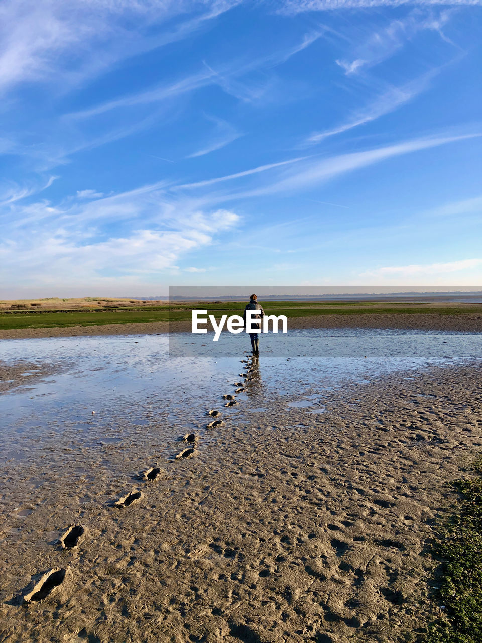Rear view of a woman leaving footprints on mudflats near the sea, under blue sky