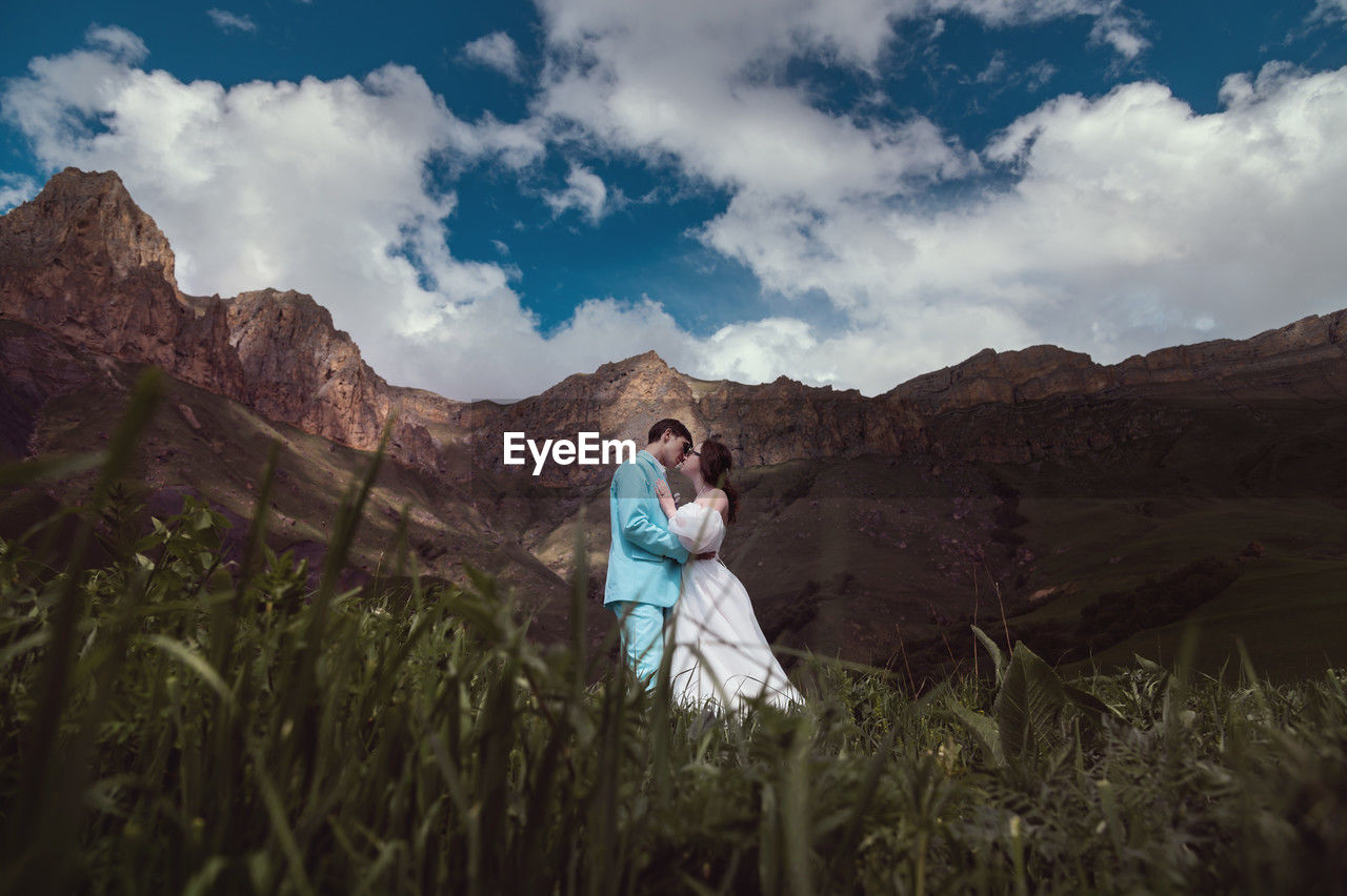 A young man and his wife stand in an embrace high in the mountains against the backdrop of epic