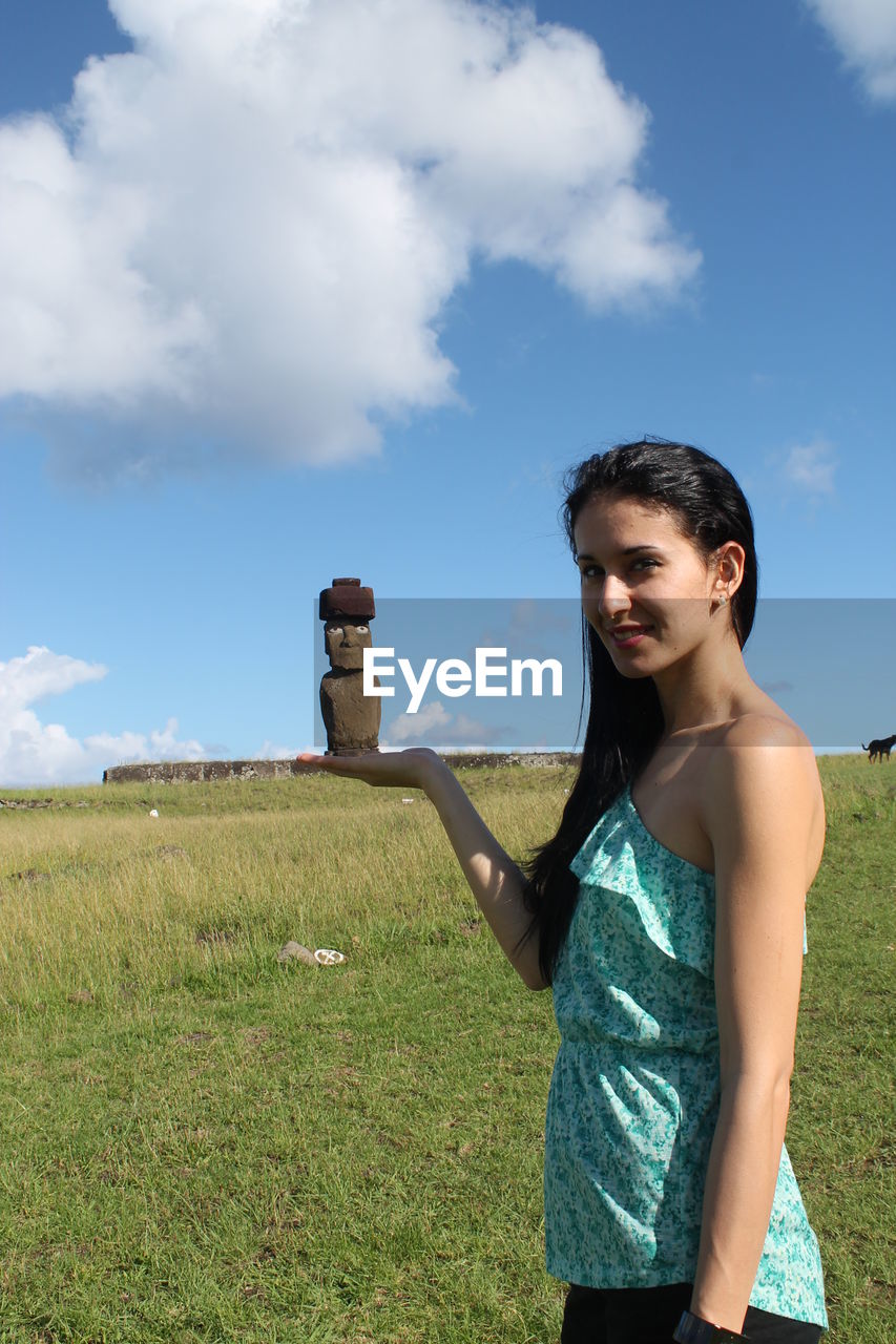 Optical illusion of young woman holding moai statue while standing on grassy field against sky