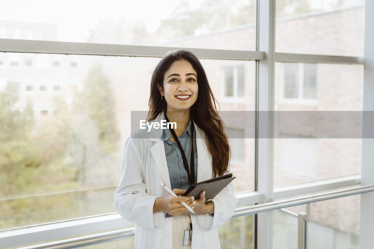 Portrait of smiling female doctor holding digital tablet and pen standing against window at hospital corridor