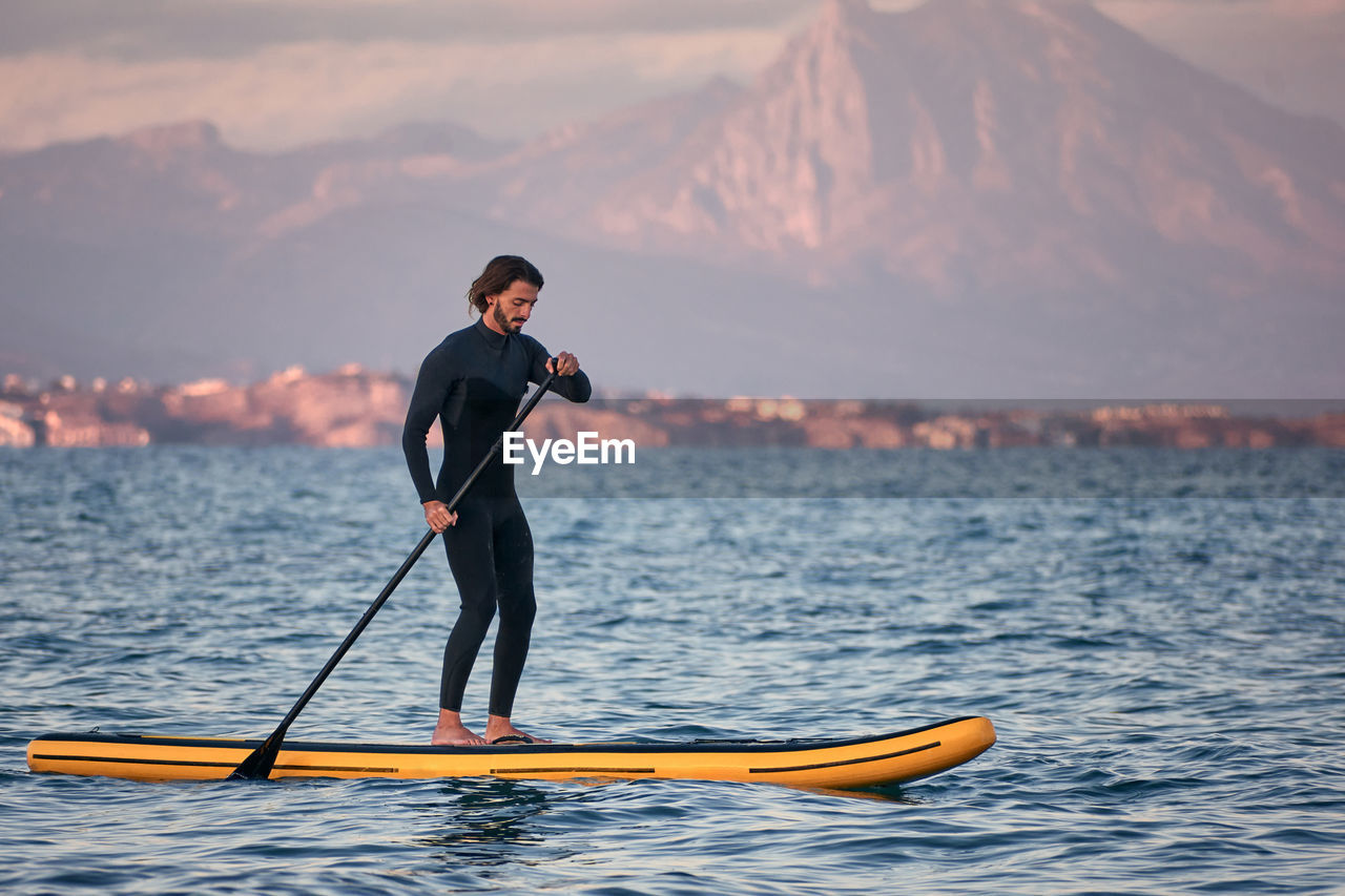 Side view of male surfer in wetsuit rowing on paddle board on sea water on background of mountains at sunset