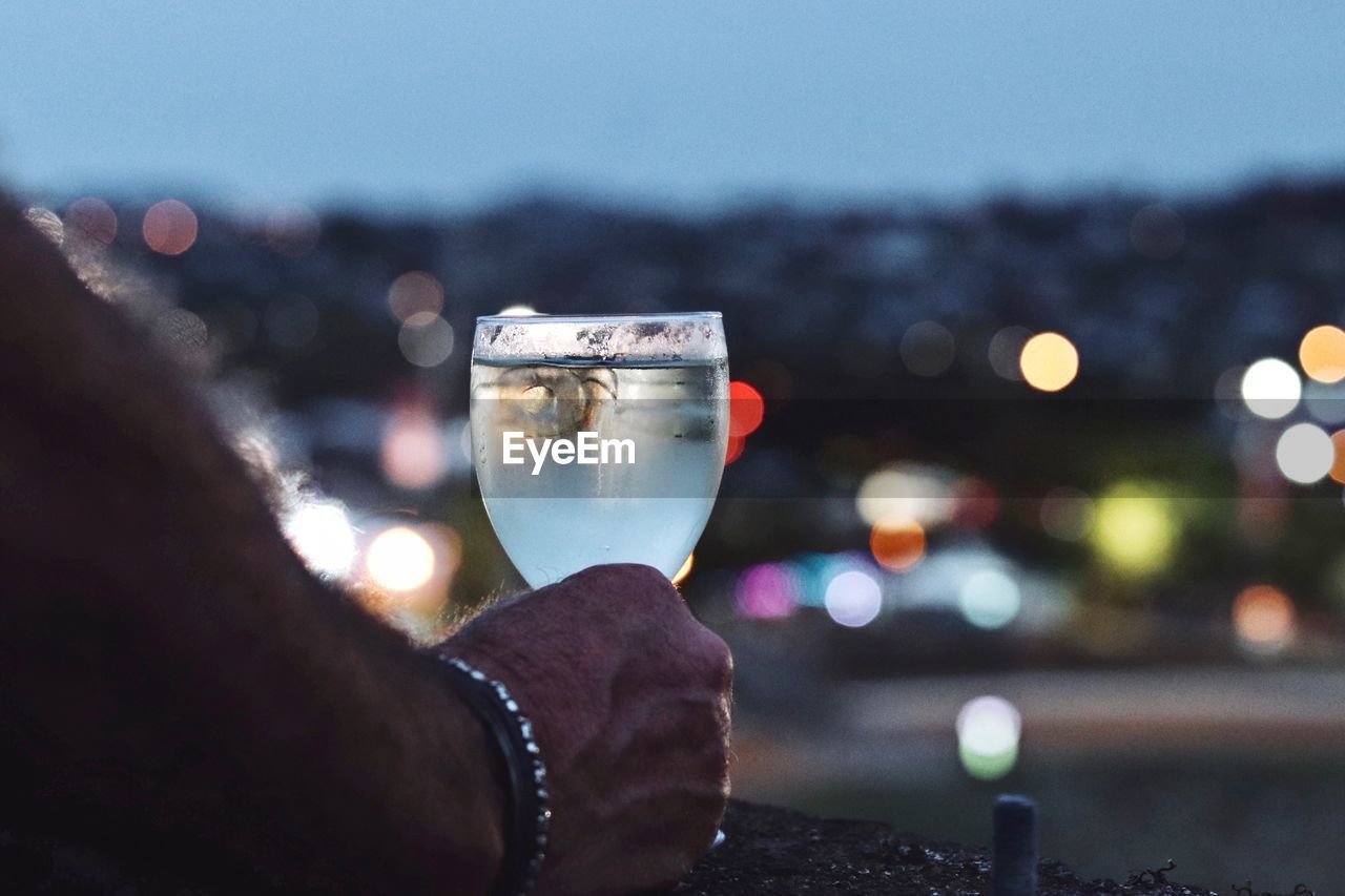 Close-up of man hand holding drink against illuminated city at night