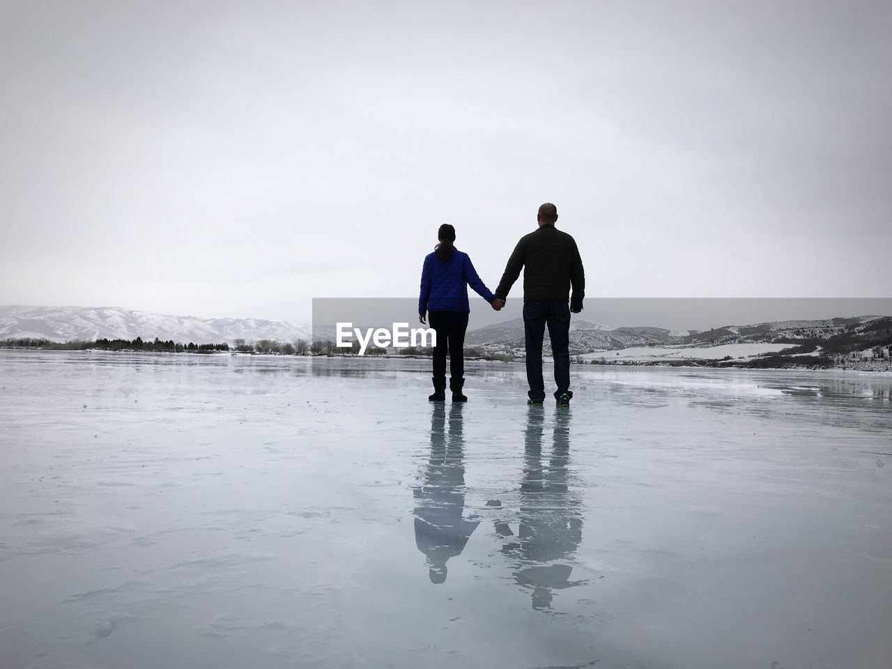 Rear view of man and woman holding hands on frozen lake against sky