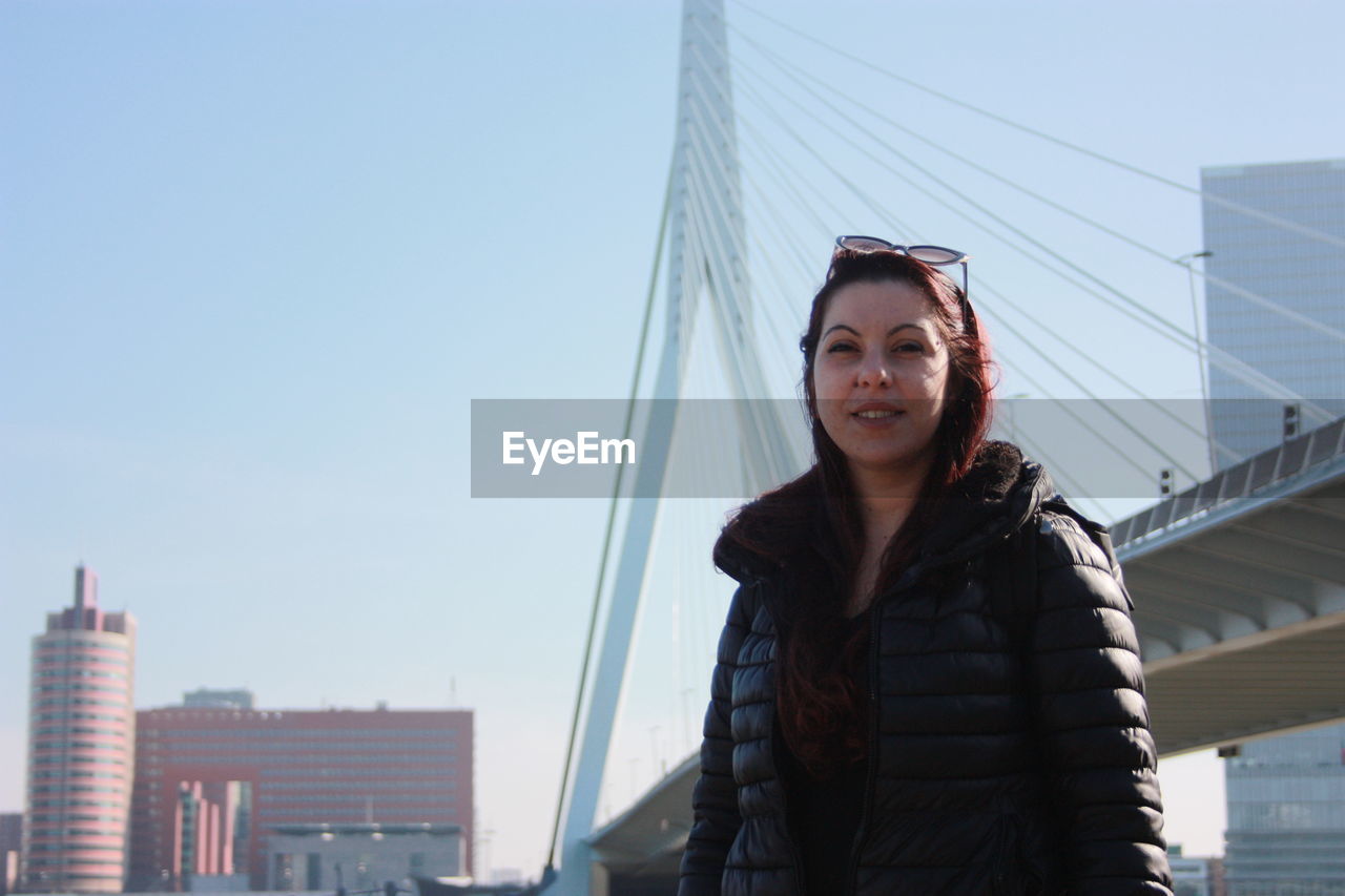 Portrait of young woman standing against bridge and sky in city