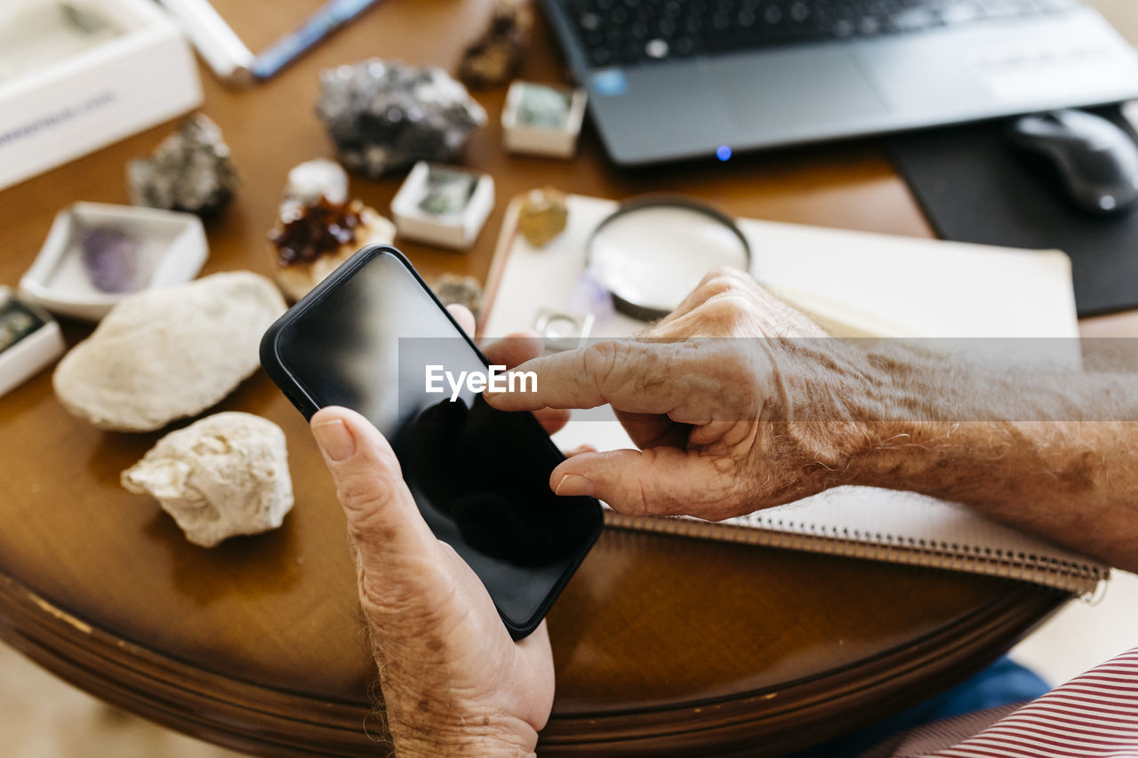 Hands of elderly retired man using smart phone while doing research on fossil and mineral at home