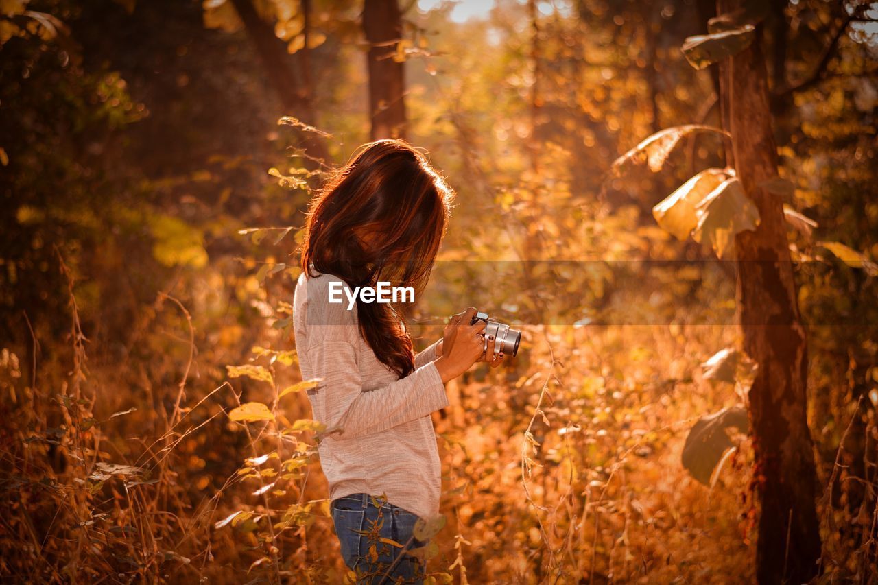 Side view of woman photographing through camera while standing at forest