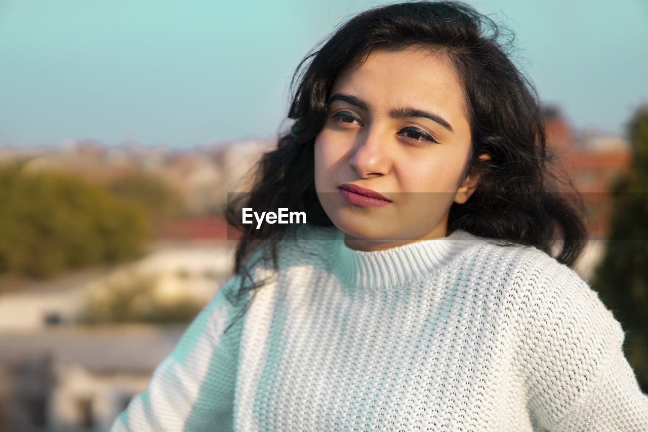 PORTRAIT OF SMILING YOUNG WOMAN STANDING AGAINST BLURRED BACKGROUND